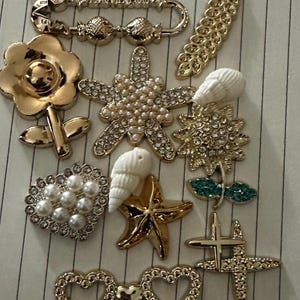 Könnte beinhalten: Eine Sammlung dekorativer Verzierungen aus Metall und Kunststoff, darunter eine Kristallblume, eine Sicherheitsnadel mit dem Text I LOVE YOU, eine goldene Blume, ein Perlen-Seestern, eine Muschel, ein Herz mit Perlen und Herzformen.