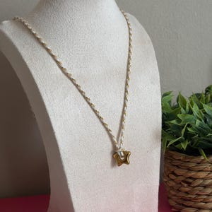 Peut inclure: Un collier composé d'un pendentif en forme d'étoile dorée suspendu à un cordon fait de fils blancs et dorés entrelacés. Le pendentif est fixé au cordon par un nœud simple, créant un design épuré et direct pour cet accessoire de bijouterie.