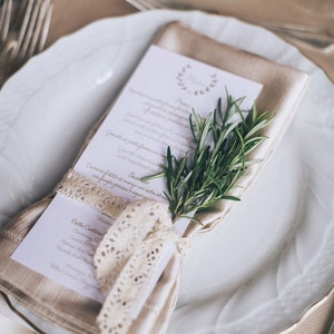 May include: A white plate with a beige napkin folded into a rectangle. The napkin is tied with a white lace ribbon and a sprig of rosemary is placed on top. The plate is set on a beige tablecloth with silverware.