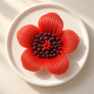 Puede incluir: Un objeto con forma de flor de color rojo brillante con cinco pétalos estriados y un centro oscuro y texturizado que presenta pequeños gránulos negros y un estambre rojo central, sobre un plato circular blanco hueso con borde elevado.