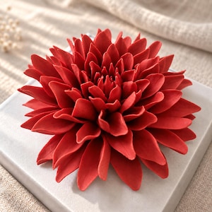 May include: A vibrant red dahlia flower sculpture with numerous pointed, layered petals radiating from the center. The flower is positioned on a smooth, square, light-colored base, showcasing a detailed, symmetrical, and multi-layered floral design.