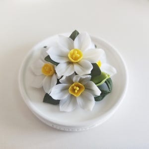 Puede incluir: Un conjunto de tres flores de narciso blancas con centros de color amarillo brillante y hojas de color verde oscuro, dispuestas sobre un pequeño plato redondo de cerámica blanca. Los pétalos están superpuestos y detallados con realismo.