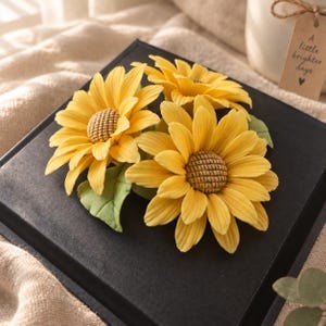 Puede incluir: Tres girasoles amarillos con centros marrones texturizados y hojas de color verde claro fijados sobre una caja negra cuadrada. Una pequeña etiqueta de papel marrón muestra el texto impreso A little brighter days y un símbolo de corazón.
