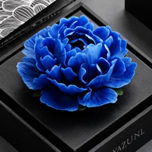 May include: A vibrant blue peony flower sculpture with layered, ruffled petals. The flower is centered on a square black box with the text YAZUNL visible on the lower right corner. The petals exhibit deep blue tones and subtle light reflections.