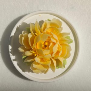 Puede incluir: Un objeto con forma de flor amarilla con pétalos en capas y pequeñas hojas verdes en la base, centrado en un plato circular blanco liso. Los pétalos están dispuestos en un patrón denso y floreciente con un centro amarillo más oscuro.