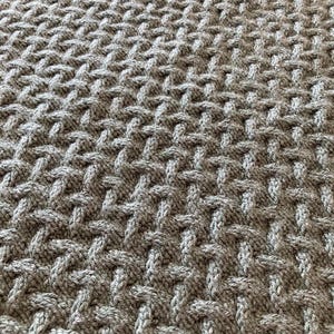 Op de afbeelding: Close-up van een grijze gebreide deken met een getextureerd, geweven patroon. Het ontwerp van de deken heeft een reeks van verweven steken, waardoor een verhoogd, tastbaar oppervlak ontstaat. De kleur is een neutraal grijs, geschikt voor diverse interieurstijlen.