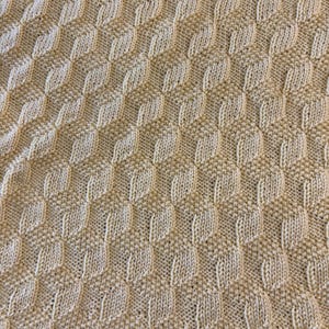 May include: A close-up of a knitted blanket with a repeating diamond pattern. The blanket is a light beige color and has a textured surface. The knitted design creates a visually interesting and tactile surface.