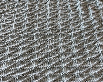 May include: Close-up of a knitted blanket in a light gray color. The blanket features a textured pattern of diagonal lines and small squares, creating a woven appearance. The soft, neutral color and intricate design suggest a cozy, handmade item.