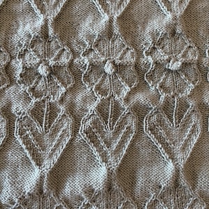 May include: Close-up of a knitted textile with a repeating pattern of stylized leaves and flowers. The fabric is a muted gray color, with the raised design creating a textured surface. The knit stitches are visible, adding to the handmade aesthetic.