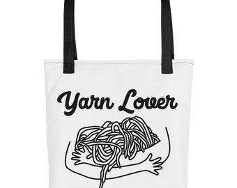 Può includere: Borsa tote bianca con manici neri e il testo "Yarn Lover" in corsivo nero. Un disegno a linea nera di una persona che abbraccia un mucchio di filato.