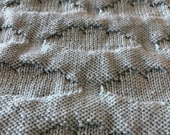May include: Close-up of a knitted blanket in a soft, muted gray color. The blanket features a textured pattern of repeating chevron shapes, creating a visually interesting surface. The knit stitches are clearly defined, showcasing the craftsmanship.