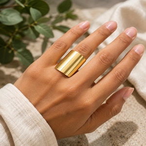 May include: A wide, polished gold-tone band ring with a smooth, cylindrical surface. The ring features a high-shine metallic finish that reflects light across its curved exterior, creating a bold and continuous band that wraps around the finger.