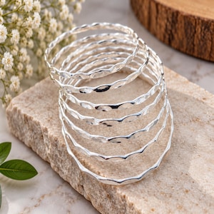 Peut inclure: Un empilement de sept bracelets joncs de couleur argentée, chacun présentant un design à bord ondulé ou festonné continu. Les bracelets sont circulaires, métalliques et possèdent une finition polie et réfléchissante qui capte la lumière.