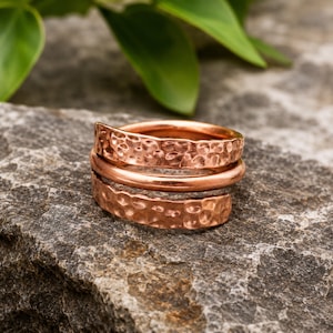 May include: A copper ring featuring a multi-layered wrap design. The band consists of three distinct sections: two outer bands with a textured, hammered finish and a smooth, polished central band, all crafted from a single piece of copper metal.