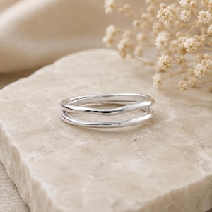 Puede incluir: Un anillo de plata pulida con un diseño de doble banda y una forma ondulada suave. El metal tiene un acabado reflectante de alto brillo, y las dos bandas están conectadas por los lados, creando una pieza de joyería singular y coherente.