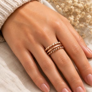 Op de afbeelding: Een set van drie roségoudkleurige ringen gedragen aan een vinger. De stapel bestaat uit één ring met een kralentextuur, één gladde band en één gedraaide ring in touwstijl. De ringen hebben een metallic afwerking en worden samen als set gedragen.