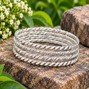 Op de afbeelding: Een set van vijf zilverkleurige armbanden, elk met een doorlopende gedraaide touwtextuur. De armbanden zijn op elkaar gestapeld en vertonen een gepolijste, reflecterende metalen afwerking die het licht opvangt over hun uniforme, cirkelvormige ontwerp.