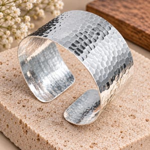 May include: A wide, open-ended cuff bracelet made of polished silver-toned metal. The exterior surface features a distinct hammered texture with numerous small, rounded indentations that reflect light across the curved, circular band design.