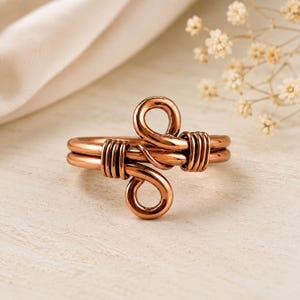 Op de afbeelding: Een koperen ring met een dubbelbandontwerp en twee overlappende lussen in het midden. De banden worden bij elkaar gehouden door strak gewikkelde koperdraad aan beide zijden van de centrale lussen, wat zorgt voor een symmetrische, gepolijste afwerking.