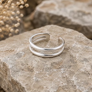 May include: A polished silver-tone open ring featuring a double-band design. The ring has a smooth, reflective surface and a gap at the back, allowing for an adjustable fit. The metal is uniform in colour and texture, showcasing a clean, rounded profile.