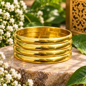 Op de afbeelding: Een stapel van vier gepolijste goudkleurige armbanden met een glad, afgerond oppervlak. De armbanden zijn cirkelvormig en hebben een reflecterende, metalen afwerking die het licht opvangt, wat zorgt voor een uniforme uitstraling.