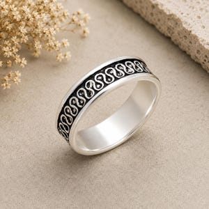May include: A silver band ring featuring a continuous, repeating S-shaped pattern engraved along the outer surface. The recessed areas of the design are darkened to provide contrast against the polished, reflective silver edges of the ring.