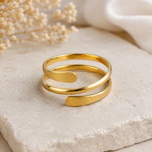Puede incluir: Un anillo de tono dorado con un diseño de bypass de dos bandas superpuestas. El metal tiene un acabado liso y pulido en la banda superior y una apariencia texturizada y martillada en la banda inferior, creando un aspecto de capas para este anillo abierto.