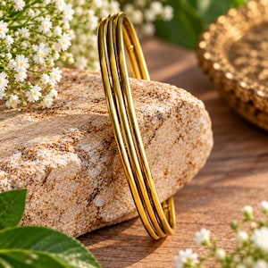 Puede incluir: Un brazalete de oro de tres bandas que presenta tres anillos circulares lisos, pulidos e interconectados. El metal tiene un acabado en oro amarillo brillante y reflectante, creando una apariencia continua y uniforme en las tres bandas apiladas.