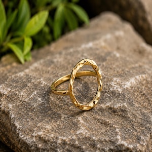 Op de afbeelding: Een goudkleurige ring met een groot, verticaal georiënteerd ovaalvormig middelpunt. Het metalen oppervlak van de ring vertoont een duidelijke gehamerde textuur, waardoor een reeks kleine, reflecterende inkepingen over de gehele ring ontstaat.