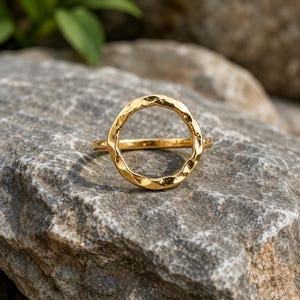 Op de afbeelding: Een gouden ring met een cirkelvormig element aan de bovenzijde met een getextureerd, gehamerd oppervlak. De ringband is dun en glad en sluit aan op de basis van de open cirkel. Het metaal heeft een reflecterende, gepolijste afwerking.