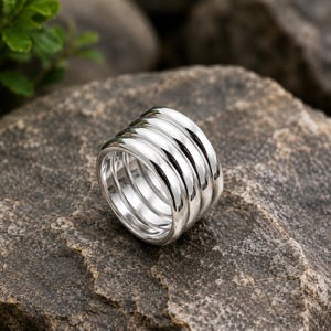 Può includere: Un ampio anello in argento con un design a fasce multiple composto da quattro anelli d'argento lisci, arrotondati e lucidati, uniti insieme per creare un unico e sostanzioso gioiello con una finitura metallica riflettente e un profilo curvo.
