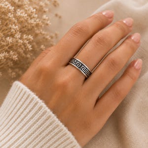 Könnte beinhalten: Ein silberfarbener Metallring mit einem durchgehenden schwarzen griechischen Mäandermuster, das in das Band eingraviert ist. Der Ring hat eine polierte Oberfläche und ein flaches Profil, das ein geometrisches Design zeigt, das den gesamten Ring umschließt.