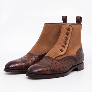 Könnte beinhalten: Ein Paar Stiefeletten mit einer dunkelbraunen, texturierten Zehenkappe mit Brogue-Details und einem Obermaterial aus beigem Wildleder. Die Stiefel haben einen seitlichen Verschluss mit sechs passenden braunen Knöpfen, einen niedrigen Absatz und eine Zugschlaufe.