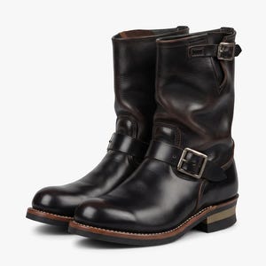 May include: A pair of dark brown leather engineer boots featuring a rounded toe, a mid-calf shaft, and two adjustable straps with silver-toned metal buckles. The boots have a stacked heel, a visible welt construction, and a dark rubber outsole.