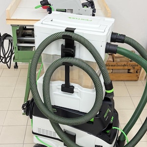 Könnte beinhalten: Ein weiß-schwarzes mobiles Festool-Absauggerät mit gestapeltem Design, einem transparenten Aufbewahrungsbehälter, einer schwarzen Schlauchhalterung und einem langen, flexiblen grün-schwarzen Saugschlauch, der außen herum gewickelt ist.