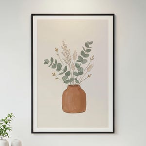 Puede incluir: Una lámina de arte enmarcada que muestra un jarrón de color terracota con un ramo de hojas de eucalipto verdes y tallos de trigo seco beige sobre un fondo crema claro, encerrado en un marco negro fino para una presentación limpia.