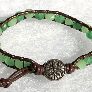 May include: Wrap bracelet featuring light green gemstone beads and brown leather cord. The bracelet has a decorative silver-tone button clasp. The beads are faceted and arranged around the leather cord.