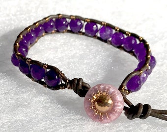 May include: A purple amethyst gemstone bracelet with a pink and gold button closure. The bracelet is made with leather cord and wire.