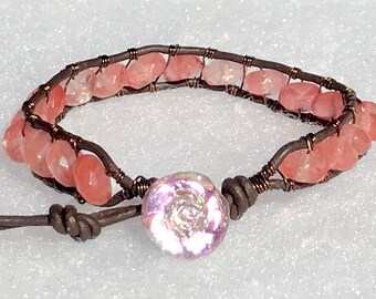 May include: A pink quartz gemstone bracelet with a brown leather cord and a pink iridescent glass bead. The bracelet is wrapped with copper wire.
