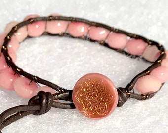May include: A wrap bracelet featuring light pink beads strung on dark brown leather. The bracelet has a decorative pink button with a gold floral design. The leather wraps around the wrist.
