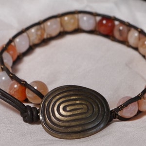 Pode incluir: Um bracelete de couro com um fecho de metal em forma de espiral e contas de ágata rosa, laranja e branca.