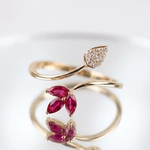 May include: A gold bypass ring featuring a leaf-shaped terminal set with small clear round diamonds and a second terminal shaped like a three-petaled flower set with marquise-cut deep red rubies, creating a delicate and open-ended design.