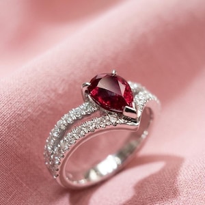 Pode incluir: Um anel de tom prateado com uma pedra preciosa vermelho-escura em forma de pera no centro. A banda tem um design em forma de V e está incrustada com pequenas pedras claras e brilhantes ao longo da haste dividida, criando um aspeto luminoso.