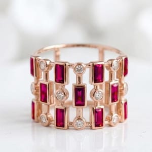 May include: A wide band ring crafted from rose-toned metal, featuring an openwork cage design. The ring is set with alternating vertical baguette-cut deep red rubies and small round brilliant-cut white diamonds arranged in a repeating pattern.