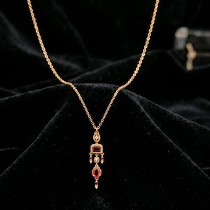 May include: A gold chain necklace featuring a pendant with a rectangular red gemstone, an oval red gemstone, and small clear accent stones. The pendant has a vertical, multi-tiered design with gold metal settings and delicate dangling elements.