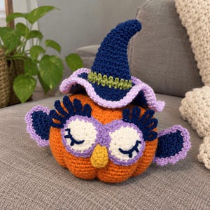 Può includere: Una zucca all'uncinetto di colore arancione con un cappello da strega blu scuro, una fascia verde chiaro e un bordo color lavanda. La zucca presenta macchie oculari bianche con ciglia blu scuro, un piccolo naso giallo e ali laterali blu scuro con bordo lavanda.
