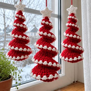 May include: Three hanging crochet Christmas tree ornaments in a spiral shape. Each tree is made of red yarn with white scalloped edges along the spiral and is topped with a small white crocheted star. They hang from red strings against a window.