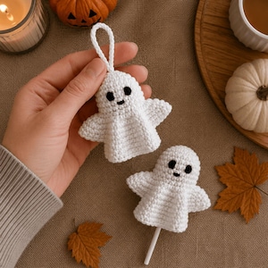May include: Two white crocheted ghost ornaments with black embroidered eyes and mouths. One ghost features a white yarn loop for hanging, while the other is attached to a white stick. Both are crafted with a textured, hand-stitched yarn finish.