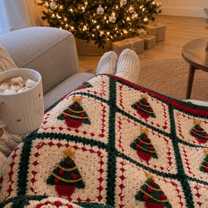 May include: A white ceramic mug with red star accents containing a hot beverage with marshmallows, resting on a cream-colored crocheted blanket featuring a repeating pattern of green Christmas trees with red ornaments and yellow stars.