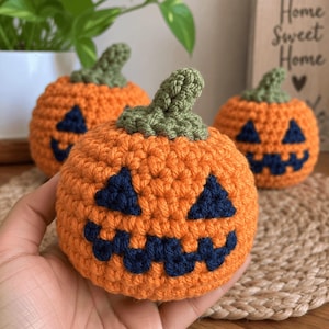 May include: Three small, round, orange crocheted pumpkins with dark blue yarn stitched into triangular eyes and jagged mouths. Each pumpkin features a textured green stem at the top. The pumpkins are crafted with a visible, chunky yarn stitch.